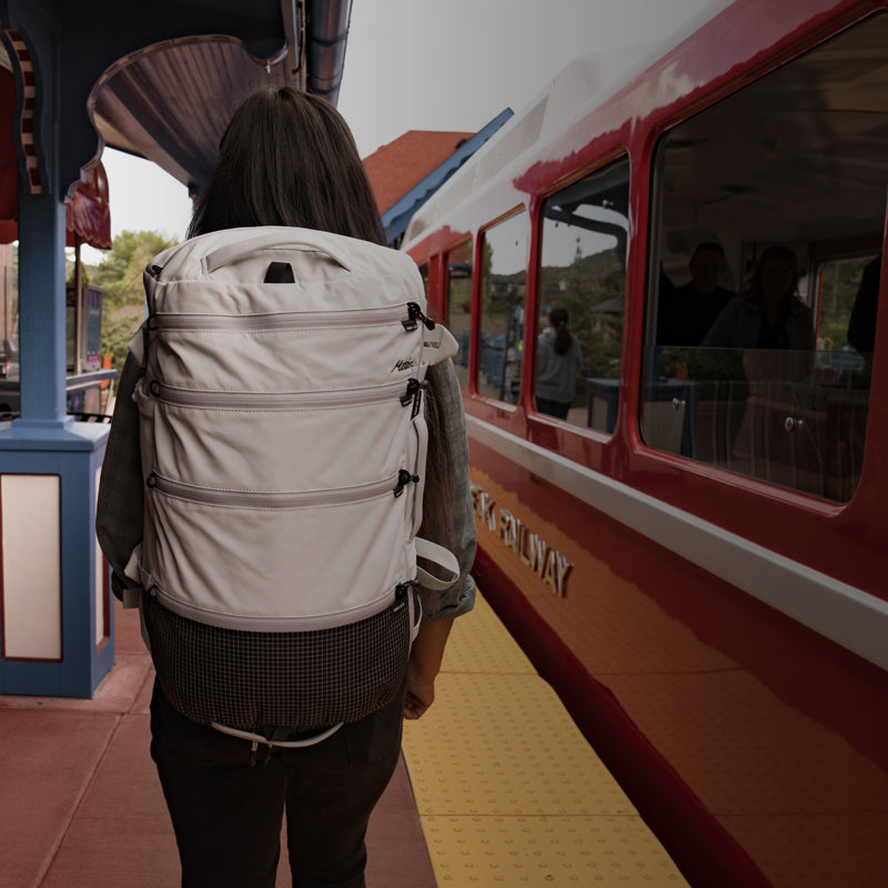 A person with a white backpack walks towards a train platform, with a colorful train in the background.